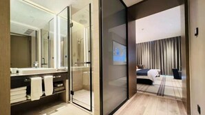 Ein modernes Hotelzimmer mit Duschkabine aus Glas, einem großen Spiegel, einem Bett mit dunklem Kopfteil und Blick auf die Stadtlandschaft durch das Fenster.