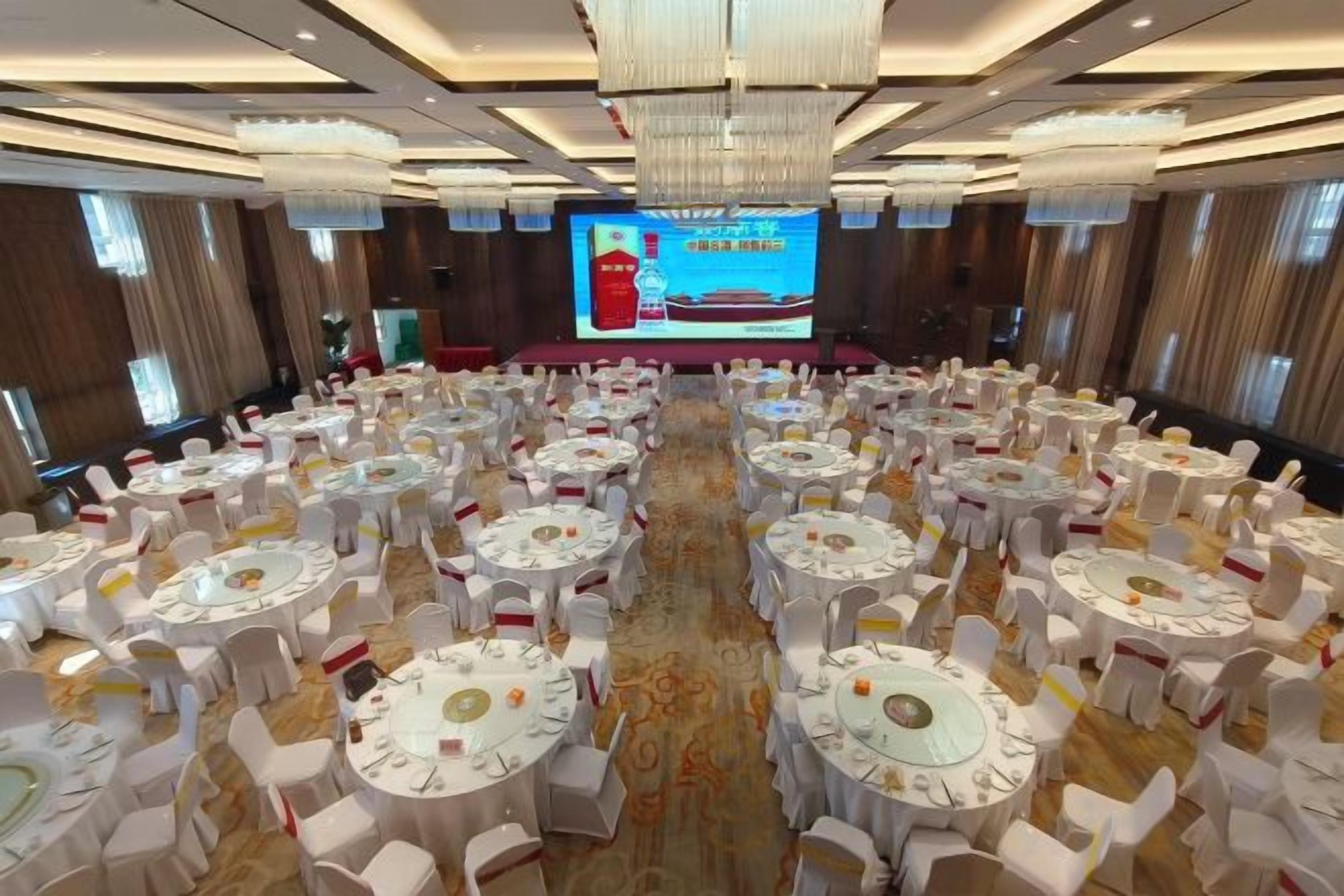 A banquet hall with round tables and chairs, a large screen at the front, and a carpeted floor.