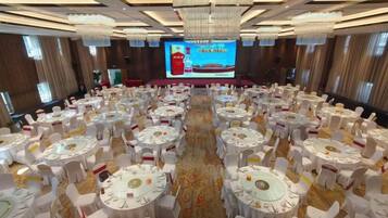A banquet hall with round tables and chairs, a large screen at the front, and a carpeted floor.