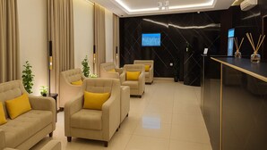 A modern hotel lobby with a row of beige sofas and armchairs, a reception desk, and a large screen displaying a crowd. - Marin Suite Al-Badiah Serviced Apartments  (Riyadh)
