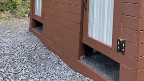 A brown wooden building with two large windows and a door, surrounded by gravel and trees. - Seneca Hideaway/Rustic Hunter’s cabin/Hot Tub (Geneva)