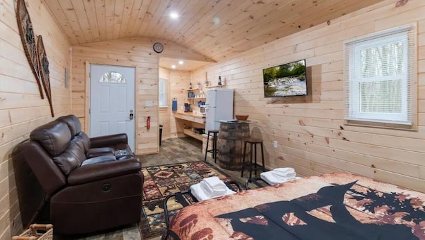 A cozy cabin with a wooden interior, a leather recliner, a bed with a patterned bedspread, a television, and a window with blinds. - Seneca Hideaway/Rustic Hunter’s cabin/Hot Tub (Geneva)