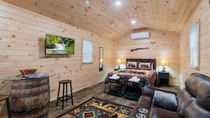 A wooden cabin interior with a bed, a leather sofa, a wooden barrel table, and a television on the wall. - Seneca Hideaway/Rustic Hunter’s cabin/Hot Tub (Geneva)