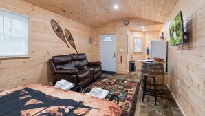 A cozy cabin room with a leather sofa, a patterned rug, a wooden barrel, and a television on the wall. - Seneca Hideaway/Rustic Hunter’s cabin/Hot Tub (Geneva)