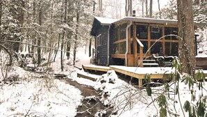 Una cabaña con terraza de madera y un pequeño porche, rodeada por un bosque nevado.