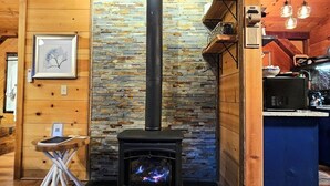 Una habitación acogedora con pared de piedra, estufa de leña negra, vigas de madera y una pequeña mesa con una silla.