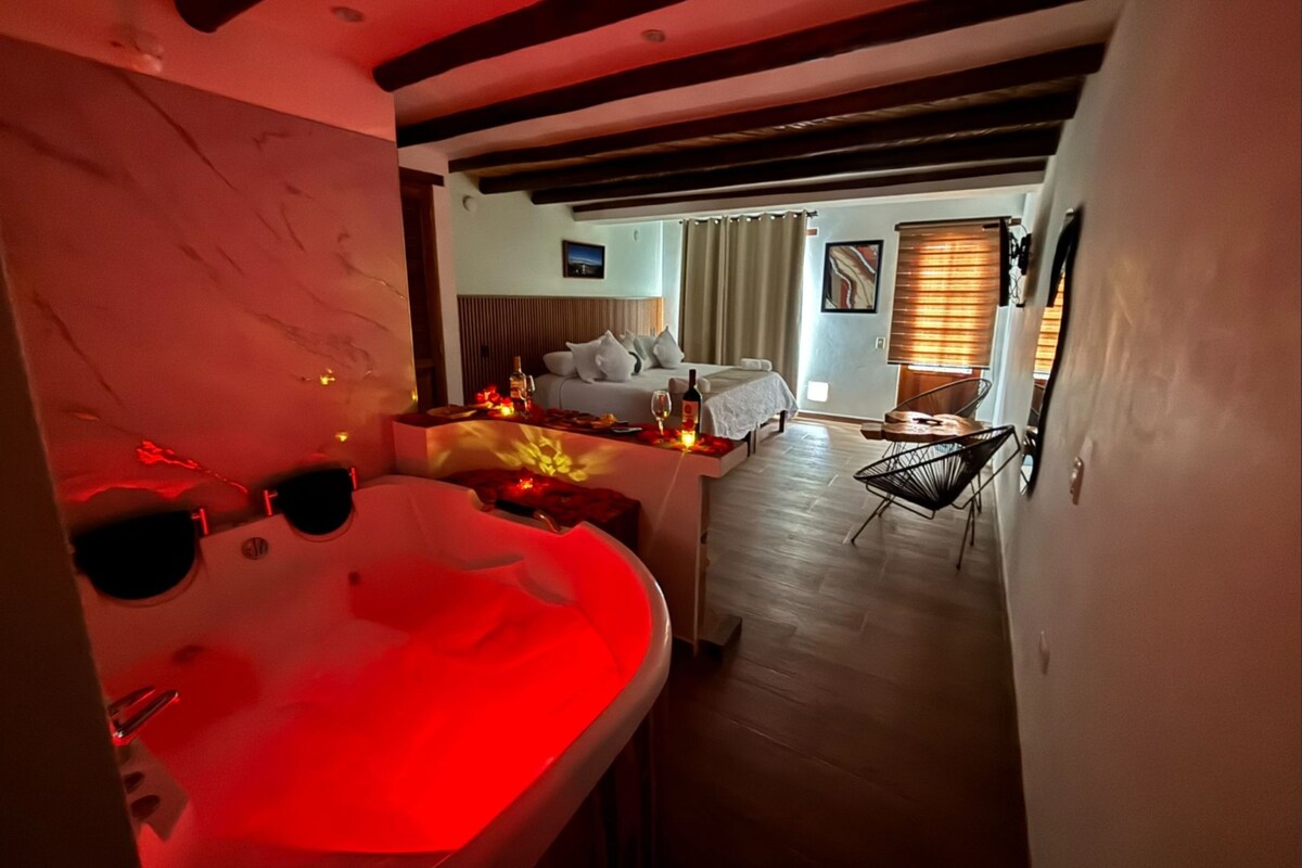 Una habitación con una cama, una pequeña mesa con velas, una silla y vistas a otra habitación con cama y cortinas.