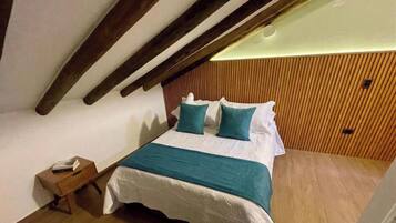 Un dormitorio con techo inclinado, vigas de madera, cama con ropa de cama blanca y almohadas color turquesa, mesita de noche con un libro y suelo de madera.