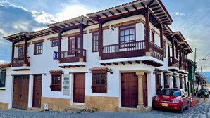 Un bâtiment à deux étages, doté d’une façade blanche et de balcons en bois, avec une enseigne bien visible indiquant qu’il s’agit d’un hôtel, et une voiture rouge garée devant.