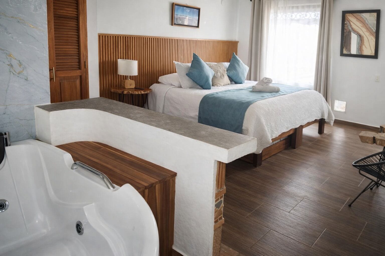 A hotel room with a wooden headboard, a bed with blue and white bedding, a bedside table with a lamp, a large bathtub, and a wooden floor.