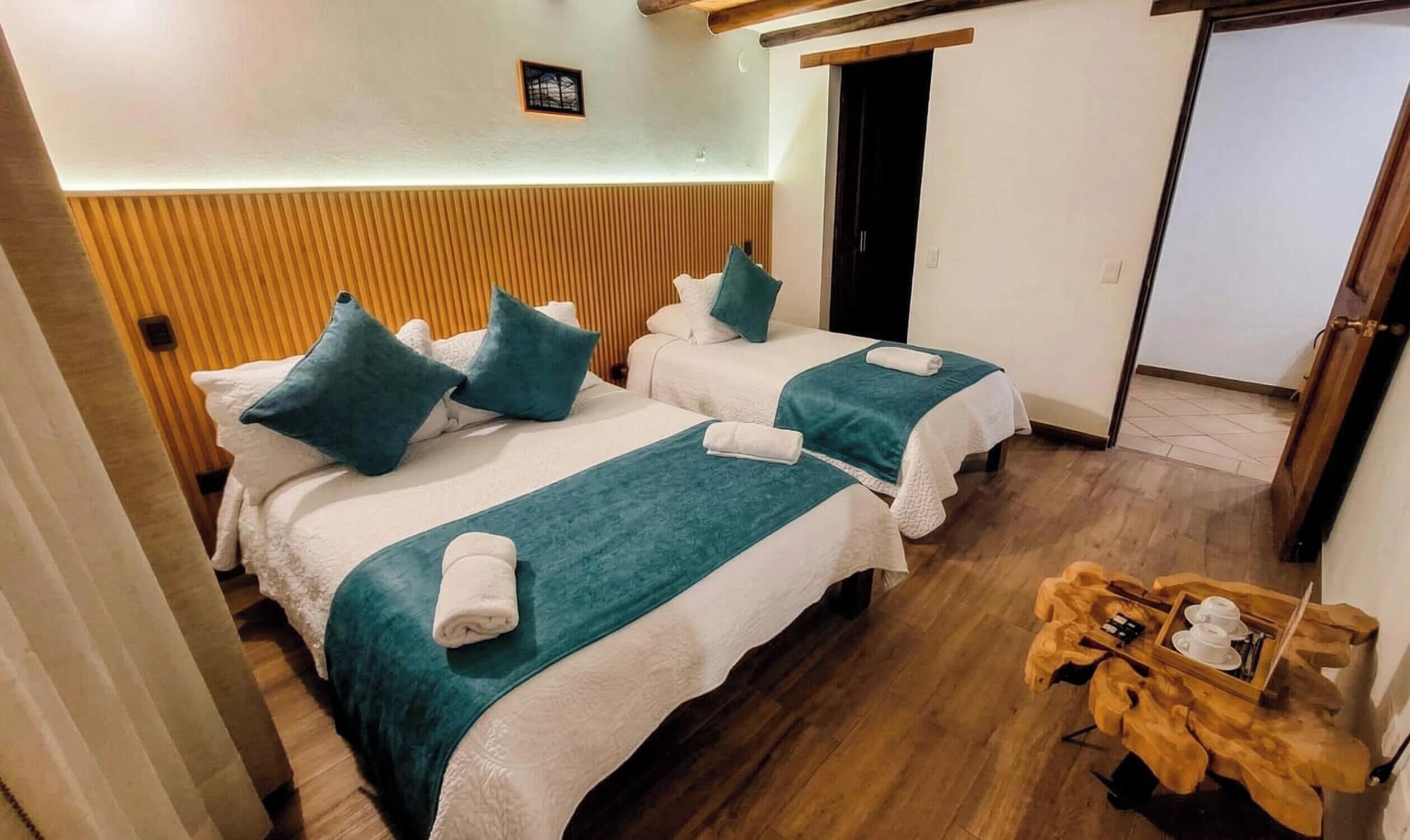 Habitación de hotel con dos camas, cada una con manta y almohadas color turquesa, cabecero de madera, una ventana pequeña y una mesita de noche con una bandeja de tazas y una caja.