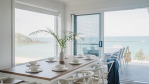A dining area with a view of the sea, a wooden table set with white plates and bowls, and white chairs. - Beach Bliss | Beachfront Luxury w/ Ocean Views (Mollymook Beach)