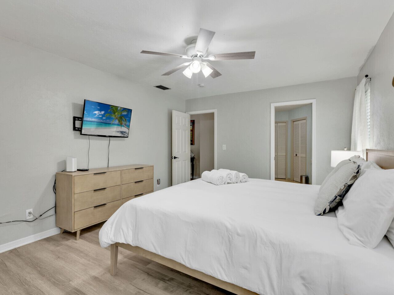 Une chambre à coucher comprenant un lit, un ventilateur de plafond, une télévision, une commode et une porte donnant sur une autre pièce.