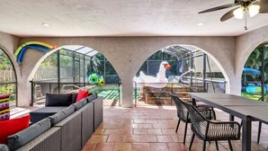 Terrassa exterior amb taula i caires de menjador, zona d'estar amb coixins, i vista a una piscina i un ànec gonflable.