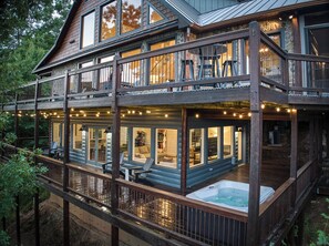 A two-story wooden cabin with a hot tub on the lower deck, large windows, and outdoor seating.