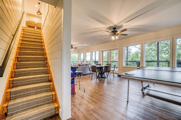 A spacious room with a large window, a ping pong table, and a staircase with a woven carpet.