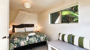 A bedroom with a bed, bedside table, lamp, and a window with a view of greenery. - Beachfront Bliss, relax in the hammock and listen to the waves on your door step (Hahei)