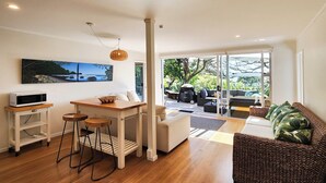 A modern living space with a kitchen area, a dining table, and a seating area with a sofa and chairs. - Beachfront Bliss, relax in the hammock and listen to the waves on your door step (Hahei)