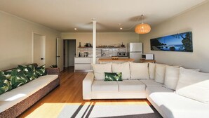 A modern living room with a large sectional sofa, a kitchen in the background, and a dining area. - Beachfront Bliss, relax in the hammock and listen to the waves on your door step (Hahei)