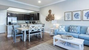 A modern living room with a blue sofa, white coffee table, and a dining area with white chairs. - D's by the Sea - 3 BR condo steps from the beach (Miramar Beach)
