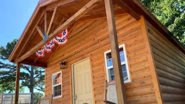 A wooden cabin with a porch, a door, and two windows. - RT 62 Motor Resort Bunkhouse (Eureka Springs)