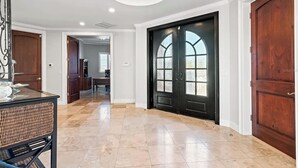 A spacious hallway with a polished tile floor, a black double door with glass panels, and a dining area with a wicker table.