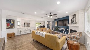 A spacious living room with a sofa, a patterned armchair, and a wooden coffee table. A kitchen with white cabinets and stainless steel appliances is visible in the background. - Cleopatra's Sunset in Ocala (Ocala)