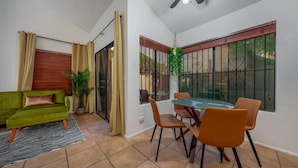 A dining area with a glass table and four chairs, a green sofa with a cushion, a potted plant, and a window with metal bars.