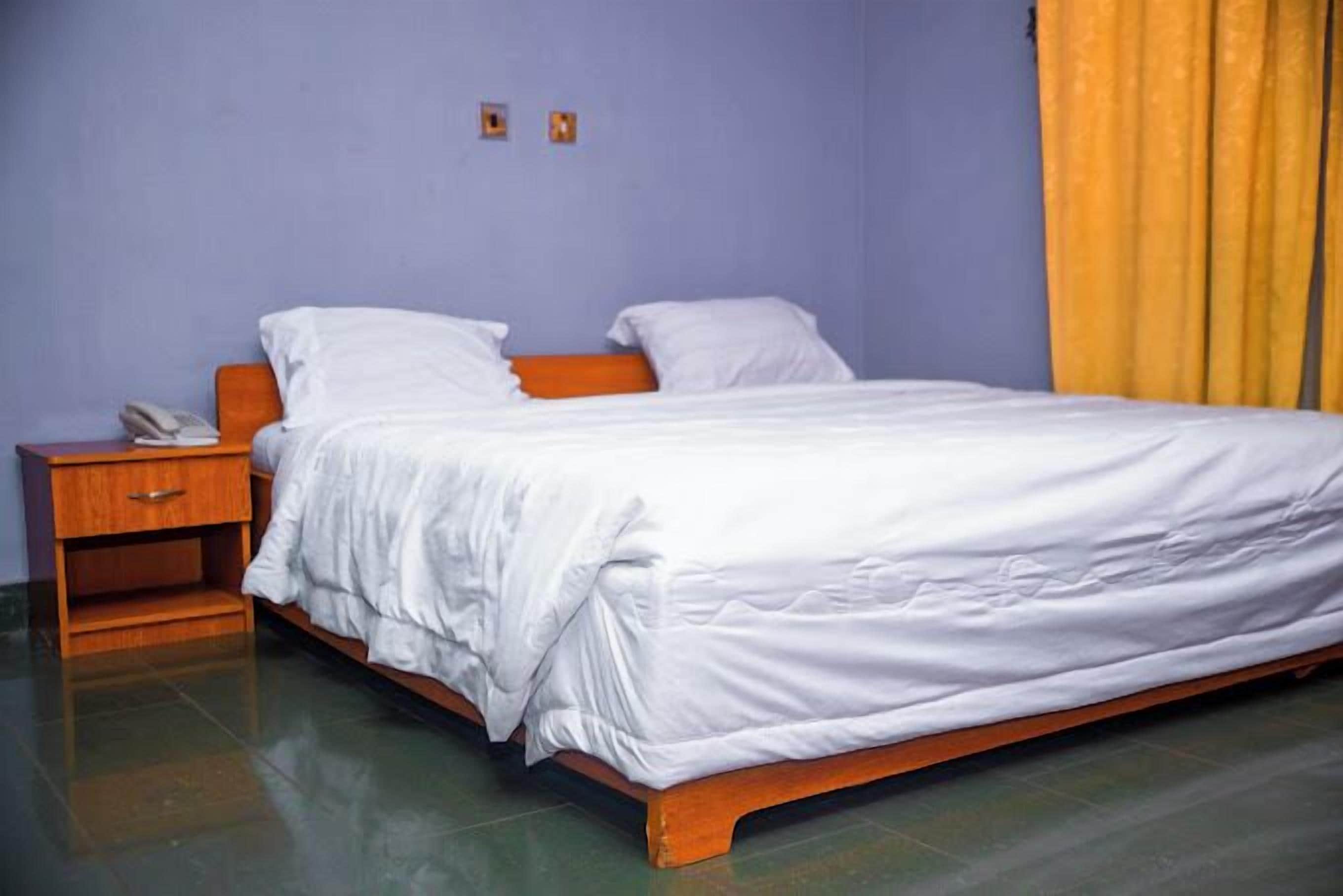 A single-bed room with a wooden frame, white bedding, a nightstand with a phone, and a yellow curtain.