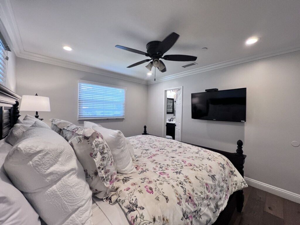 Ein Schlafzimmer mit einem Bett, einem Deckenventilator, einem an der Wand montierten Fernseher und einem Fenster mit Jalousien.