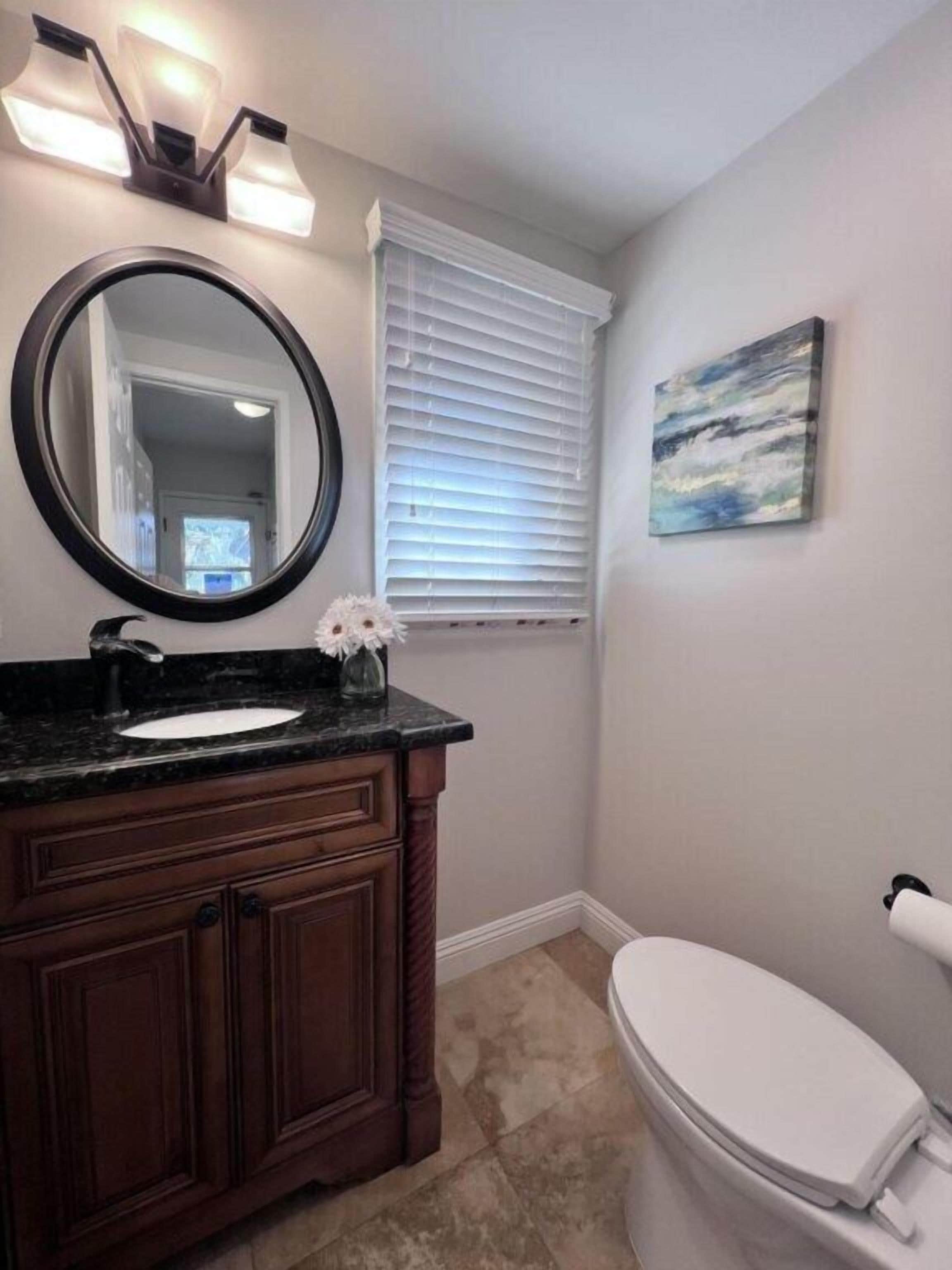 Une salle de bain avec un miroir rond, un lavabo, des toilettes, une fenêtre avec des stores et un tableau au mur.