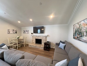 A living room with a fireplace, a television, a sofa, a dining table with chairs, and artwork on the walls.
