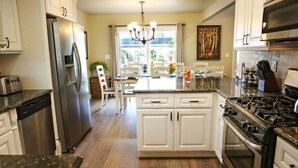 Une cuisine avec des meubles blancs, un réfrigérateur en acier inoxydable, une cuisinière à gaz et un coin repas agrémenté d’un lustre.