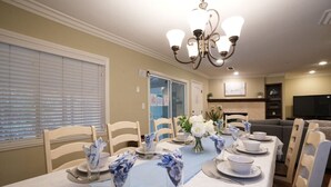 Une salle à manger avec une longue table dressée pour six personnes, des chaises, un lustre et un téléviseur en arrière-plan.