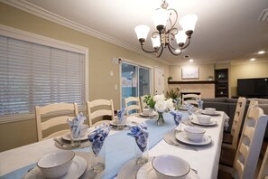 A dining room with a long table set for six, chairs, a chandelier, and a television in the background.