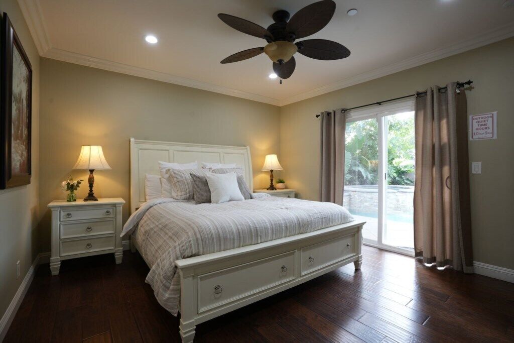 A bedroom with a bed, two bedside tables with lamps, a ceiling fan, and a sliding glass door to the outside.