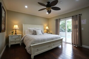 Une chambre à coucher avec un lit, deux tables de chevet avec des lampes, un ventilateur de plafond et une porte-fenêtre coulissante donnant sur l’extérieur.