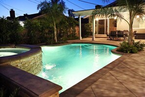 Ein Schwimmbecken mit Steinkante, umgeben von einer Terrassenfläche mit Stühlen und einer überdachten Sitzgelegenheit.