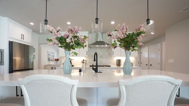 Una cocina moderna con muebles blancos, una isla central con dos sillas y jarrones decorativos con flores.