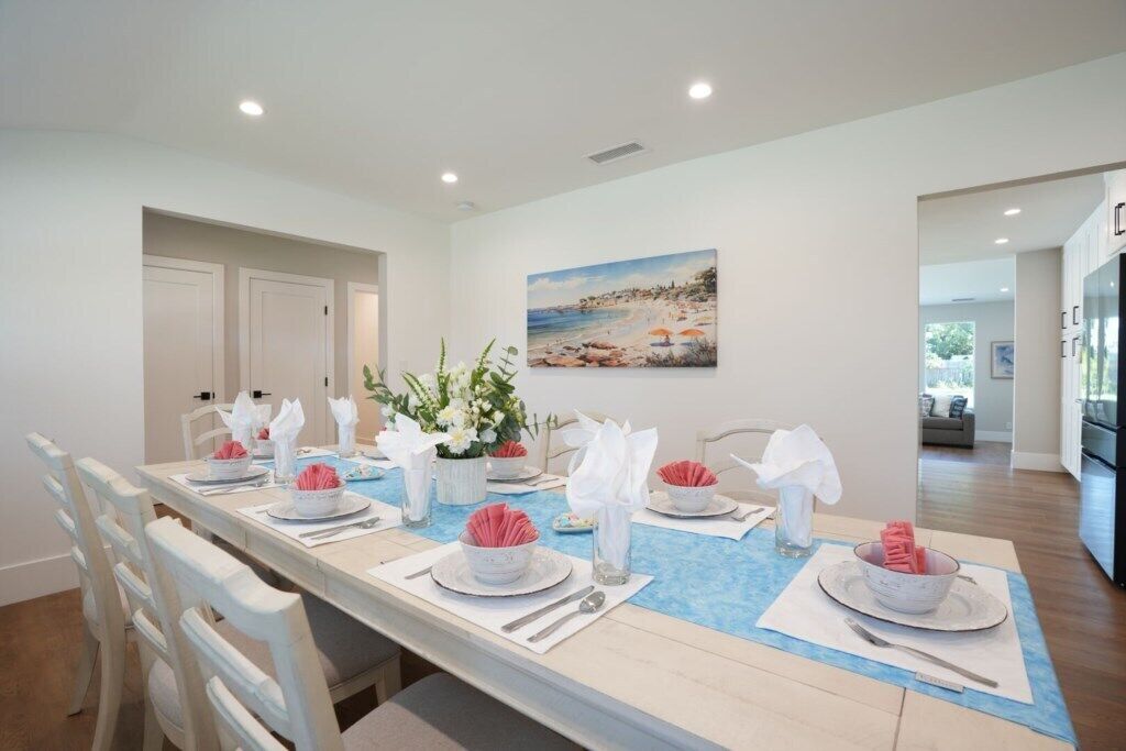 Un coin repas avec une longue table dressée pour six personnes, des chaises blanches, un chemin de table bleu et un tableau représentant une scène de plage accroché au mur.