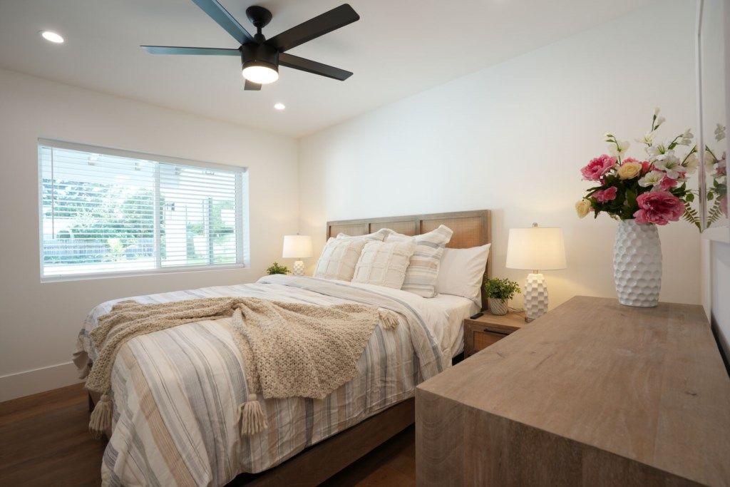 A bedroom with a bed, bedside lamps, a nightstand, a wooden desk, a ceiling fan, and a window with blinds.