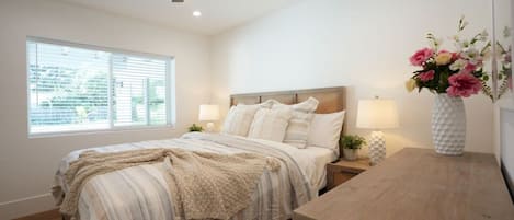 A bedroom with a bed, bedside lamps, a nightstand, a wooden desk, a ceiling fan, and a window with blinds.
