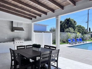 Um pátio externo com mesa, cadeiras, churrasqueira e área de piscina com espreguiçadeiras.