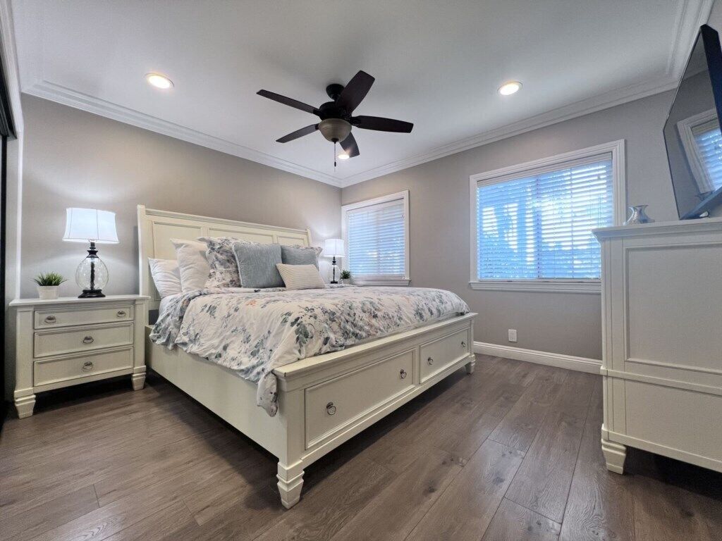 Ein Schlafzimmer mit Bett, Nachttischen, Kommode, Deckenventilator und Fenstern mit Jalousien.