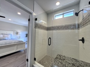 A bathroom with a glass shower enclosure, a tiled shower area, and a visible bed in the adjacent room.