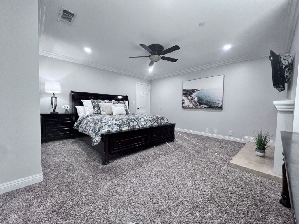 A bedroom with a bed, a ceiling fan, a nightstand, a lamp, a framed picture on the wall, and a carpeted floor.