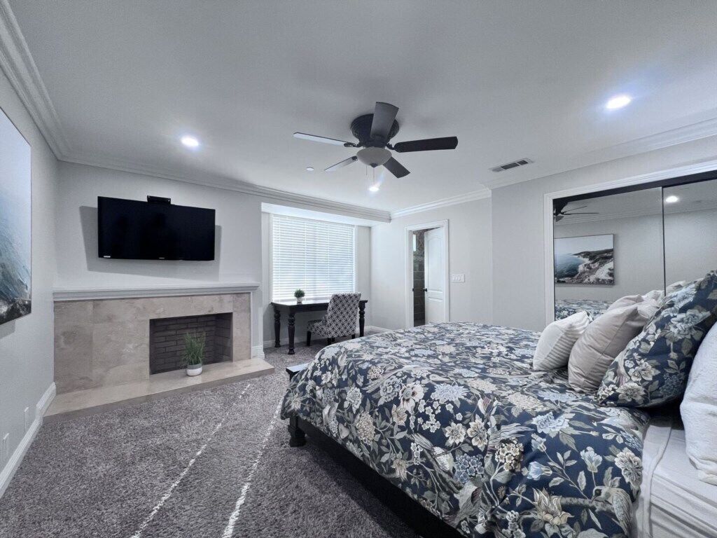 A bedroom with a bed, a ceiling fan, a TV mounted on the wall, a fireplace, and a window with blinds.