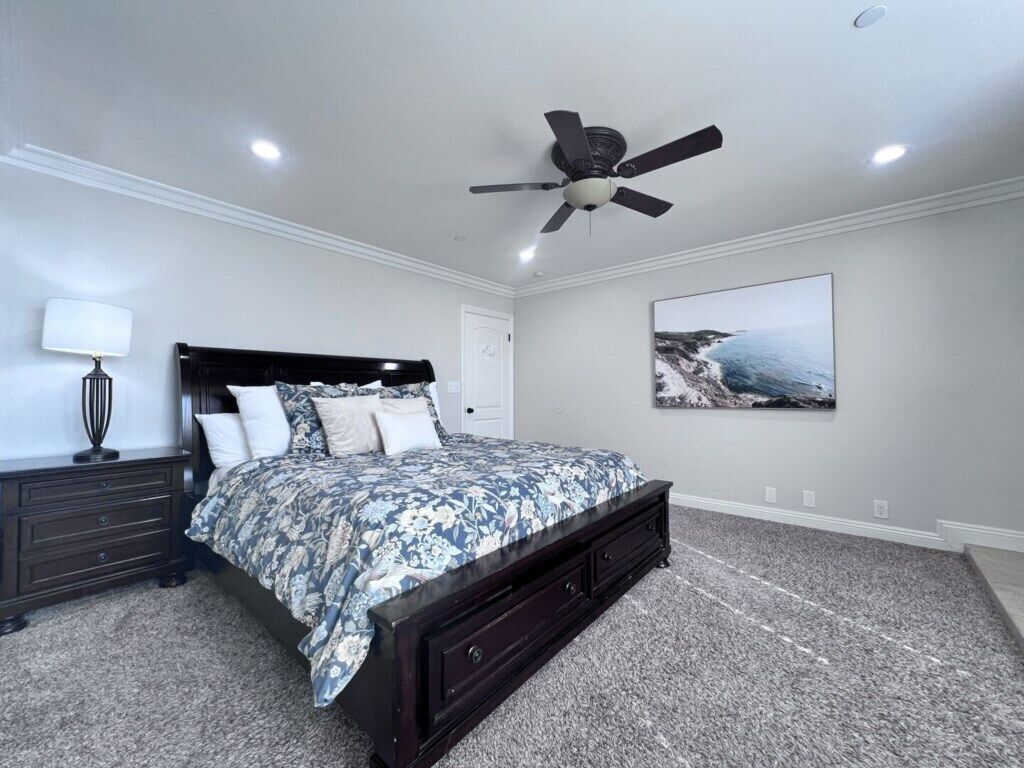 A bedroom with a bed, a ceiling fan, a nightstand, a lamp, and a painting on the wall.