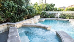 A swimming pool with a waterfall feature, surrounded by lush greenery and a white fence. - Sensational 6 Bedroom 4 Bathroom Pool Home (3 Master Bedrooms W/bathrooms) (Anaheim)
