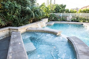 A swimming pool with a waterfall feature, surrounded by lush greenery and a white fence.